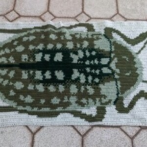 May include: A crocheted rug featuring a green and white beetle design. The rug is made with a white yarn and has a green beetle with black accents in the center.