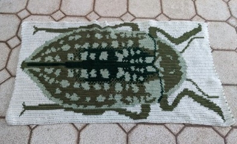 May include: A crocheted rug featuring a green and white beetle design. The rug is made with a white yarn and has a green beetle with black accents in the center.