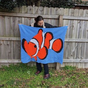 May include: A blue crocheted blanket with a large orange and white clownfish design. The blanket is being held up by a person.