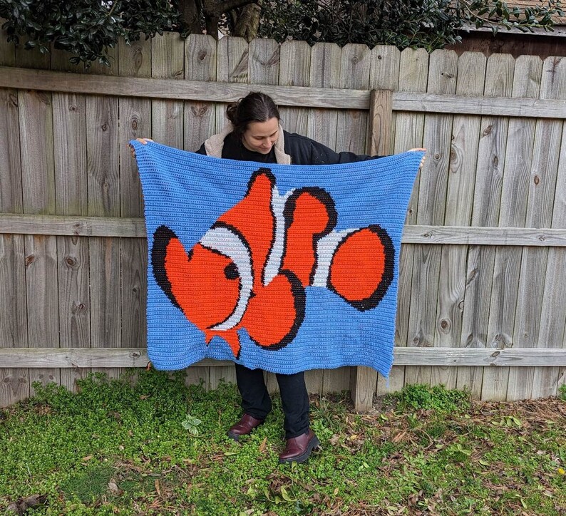 May include: A blue crocheted blanket with a large orange and white clownfish design. The blanket is being held up by a person.