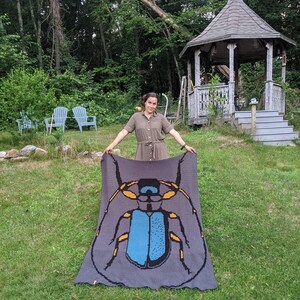 May include: A woman holds up a crocheted blanket featuring a large blue and yellow beetle design. The blanket is gray with a dark gray border.