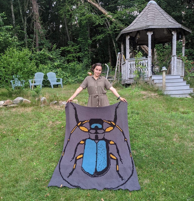 May include: A woman holds up a crocheted blanket featuring a large blue and yellow beetle design. The blanket is gray with a dark gray border.