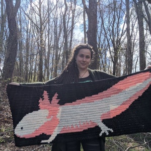 May include: A person holding a crocheted blanket featuring a pink and white axolotl. The blanket is black with a white and pink axolotl design.