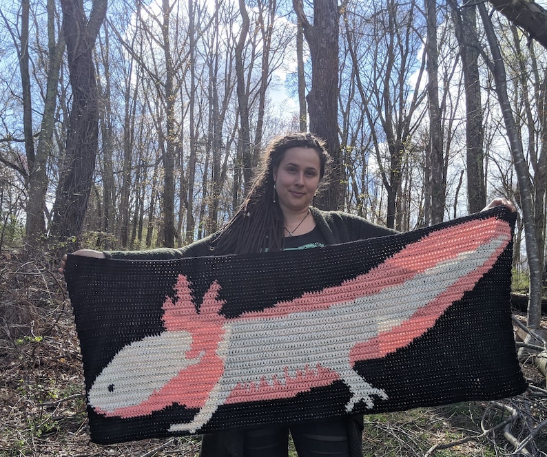 May include: A person holding a crocheted blanket featuring a pink and white axolotl. The blanket is black with a white and pink axolotl design.