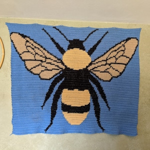 May include: A crocheted wall hanging featuring a bee with black and brown wings and a yellow and black body on a blue background.