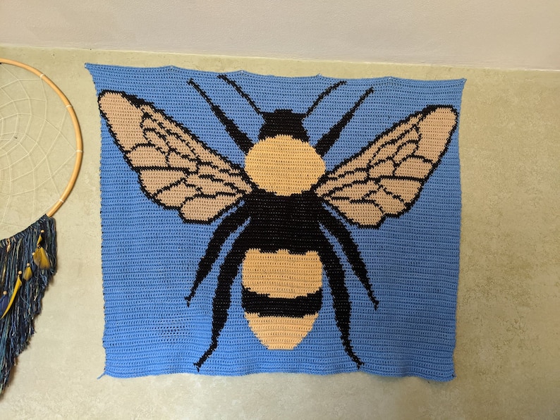 May include: A crocheted wall hanging featuring a bee with black and brown wings and a yellow and black body on a blue background.