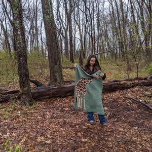 May include: A person in a woodland setting holds up a green crocheted blanket featuring a giraffe design. The blanket is crafted using brown, tan, and white yarn.