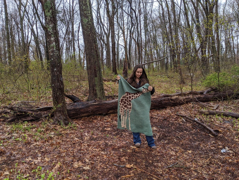 May include: A person in a woodland setting holds up a green crocheted blanket featuring a giraffe design. The blanket is crafted using brown, tan, and white yarn.