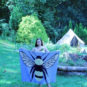 May include: A woman holds a crocheted blanket featuring a large bee design. The blanket is purple with yellow and black accents. The woman is standing in a grassy area with a tent in the background.