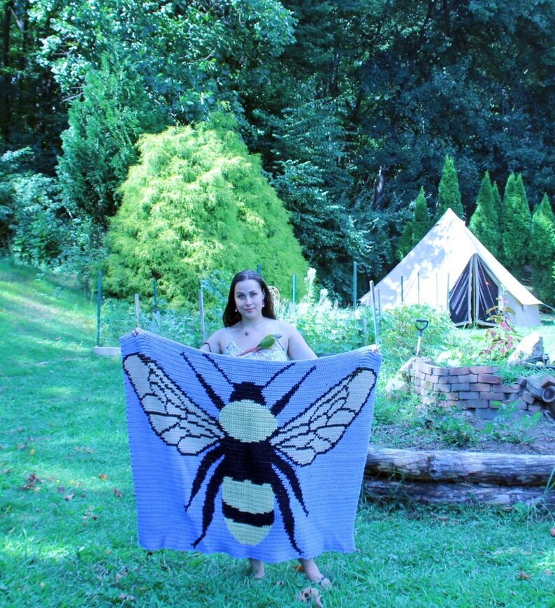 May include: A woman holds a crocheted blanket featuring a large bee design. The blanket is purple with yellow and black accents. The woman is standing in a grassy area with a tent in the background.
