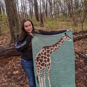 May include: A woman is holding up a crocheted blanket featuring a giraffe design. The blanket is teal blue with brown and beige accents. The woman is wearing a black long-sleeved shirt with the word "AGAINST" printed on it and blue jeans.