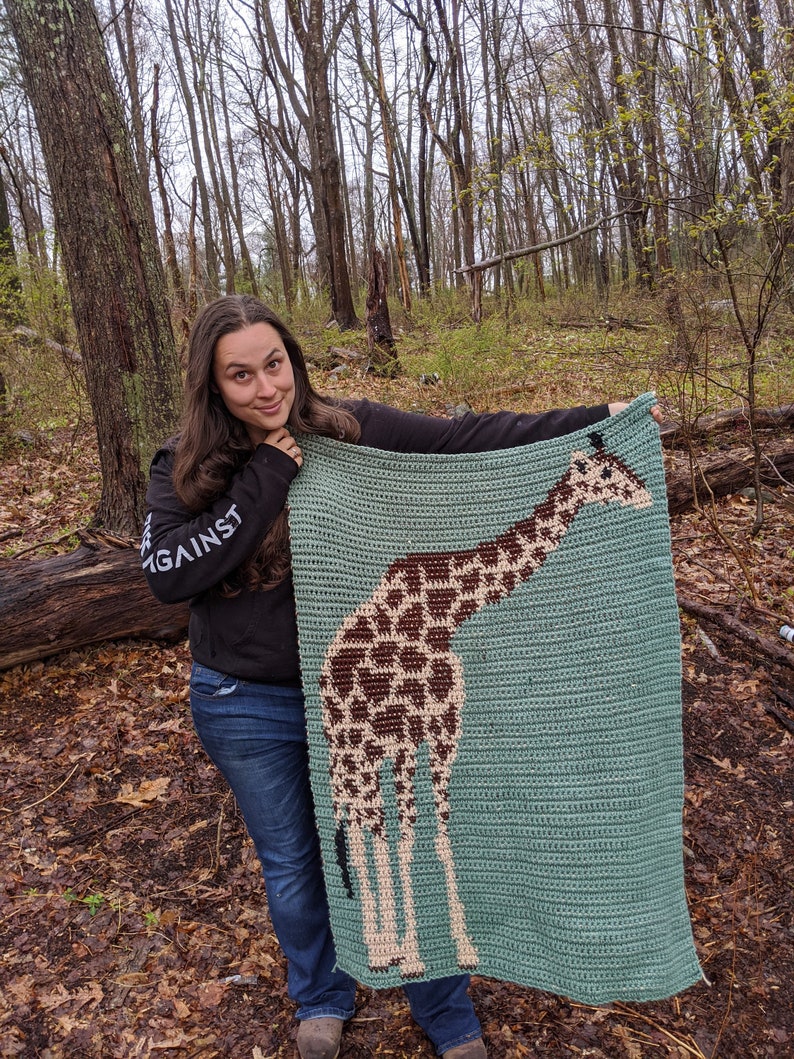 May include: A woman is holding up a crocheted blanket featuring a giraffe design. The blanket is teal blue with brown and beige accents. The woman is wearing a black long-sleeved shirt with the word "AGAINST" printed on it and blue jeans.