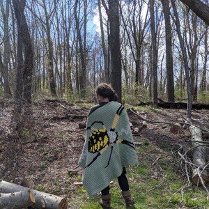 May include: A person wearing a green crocheted blanket with a yellow and black butterfly design. The blanket is worn over a black shirt and pants. The person is standing in a forest setting.