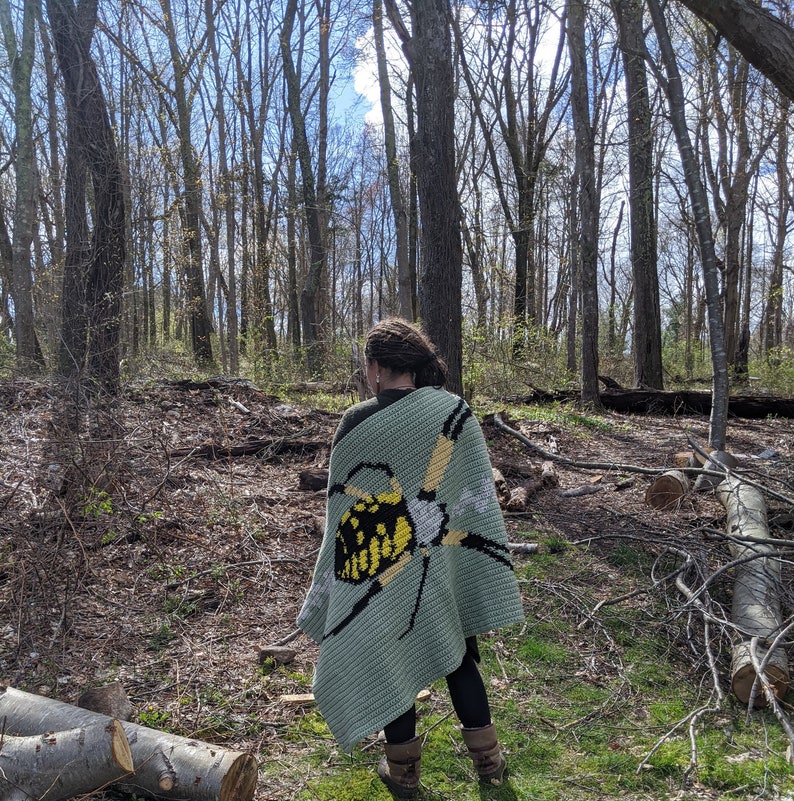 May include: A person wearing a green crocheted blanket with a yellow and black butterfly design. The blanket is worn over a black shirt and pants. The person is standing in a forest setting.