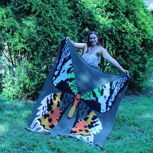 May include: A green crochet blanket with a large butterfly design in shades of orange, white, green, and black. The blanket is being held up by a person.