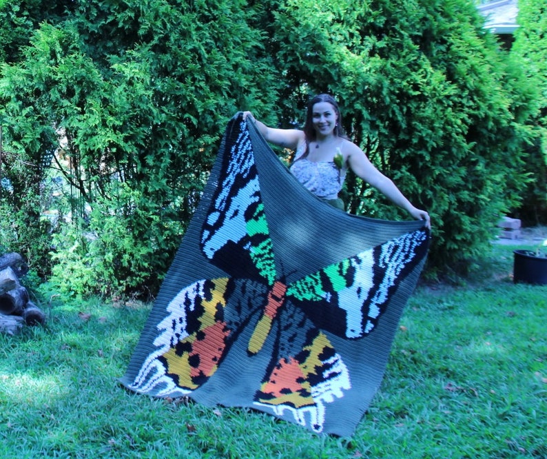 May include: A green crochet blanket with a large butterfly design in shades of orange, white, green, and black. The blanket is being held up by a person.