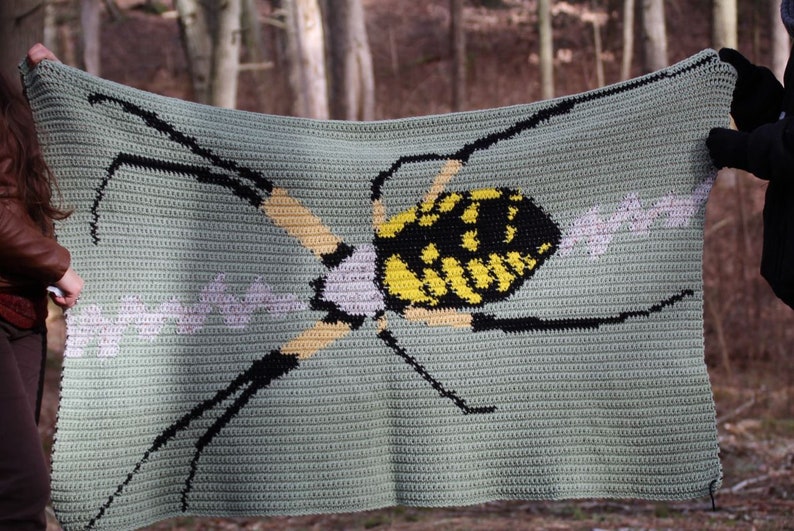 May include: A crocheted blanket featuring a yellow and black spider with a white web design. The blanket is green and has a textured look.
