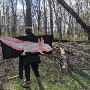 May include: A person wearing a black jacket and jeans is holding a crocheted blanket featuring a pink and white axolotl on a black background.