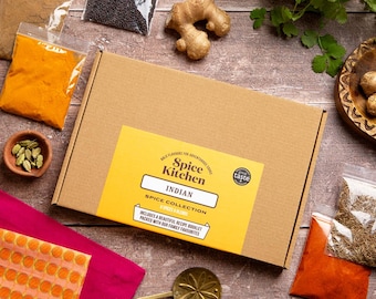 May include: A rectangular brown cardboard box labeled Spice Kitchen Indian Spice Collection with a yellow sticker. The box is surrounded by various spices in clear plastic bags, a ginger root, green cardamom pods, and a gold metal spice spoon.