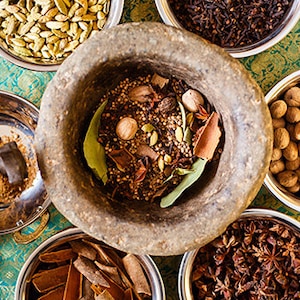May include: A collection of spices in bowls, including cardamom pods, cloves, star anise, nutmeg, and bay leaves. The spices are arranged on a patterned surface and are ready to be ground in a mortar and pestle.