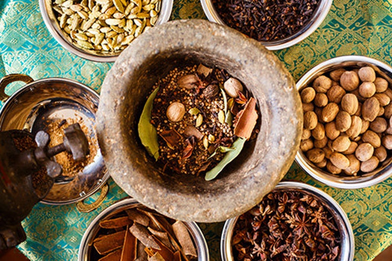 May include: A collection of spices in bowls, including cardamom pods, cloves, star anise, nutmeg, and bay leaves. The spices are arranged on a patterned surface and are ready to be ground in a mortar and pestle.