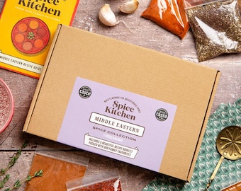 May include: A rectangular cardboard box labeled Spice Kitchen Middle Eastern Spice Collection with a purple sticker, accompanied by a yellow recipe booklet, several clear plastic bags containing various ground spices, garlic cloves, and a gold spoon.
