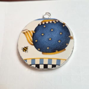 May include: A circular pendant featuring a blue teapot with yellow polka dots, a yellow striped handle, and a small bee illustration. The base of the design includes a blue, white, and black checkered pattern. A silver metal jump ring is attached to the top.