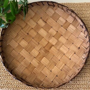 May include: Round woven bamboo tray with a tightly woven pattern. The tray has a dark brown border and is set on a textured, light brown surface. Green leaves are visible in the upper left corner. This tray is ideal for serving food or as a decorative piece.