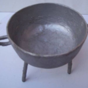 May include: A small, gray metal cauldron with a rounded bowl, two handles, and three short legs. The cauldron has a simple, utilitarian design, suitable for cooking or decorative purposes.