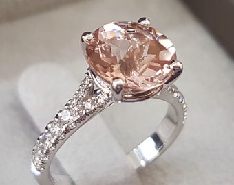 Op de afbeelding: Een delicate verlovingsring met een grote, ronde, lichtroze edelsteen omringd door een band van kleinere, glinsterende diamanten.