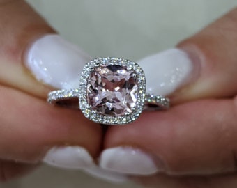 Könnte beinhalten: Ein Ring mit einem großen, kissenförmigen, blassrosa Edelstein, der von einem Heiligenschein aus kleineren Diamanten umgeben ist. Die Ringschiene ist ebenfalls mit kleineren Diamanten besetzt.