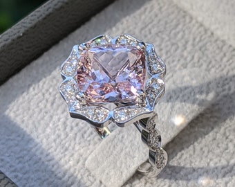 Könnte beinhalten: Ein zarter Ring mit einem großen, quadratischen rosafarbenen Edelstein, der von kleineren, funkelnden Diamanten umgeben ist. Der Ring hat ein florales Design mit filigranen Details.