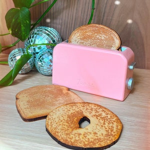 May include: A pink toy toaster with two wooden slices of bread, one whole and one with a hole in the middle, sitting on a wooden surface.