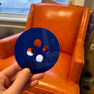May include: A blue plastic disc with four holes in the center. The disc is being held in a hand in front of an orange chair.