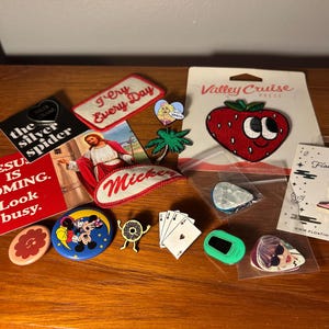 May include: A collection of assorted enamel pins, patches, and stickers featuring various designs including a strawberry, a cowboy boot, playing cards, a donut, Mickey Mouse, and text-based graphics like Jesus is Coming and I Cry Every Day.