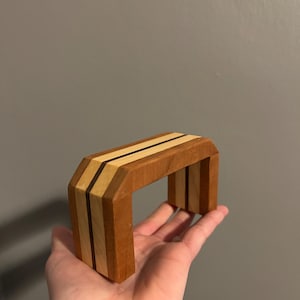 May include: A small, wooden arch-shaped object with a geometric design. The arch is constructed from various wood tones, including light and dark brown, and beige. The top features two thin, dark brown stripes. The object is held in a hand.