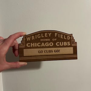 May include: Wooden sign with the text "WRIGLEY FIELD HOME OF CHICAGO CUBS" and "GO CUBS GO!" in a decorative font. The sign is rectangular with a curved top and is held by a hand.
