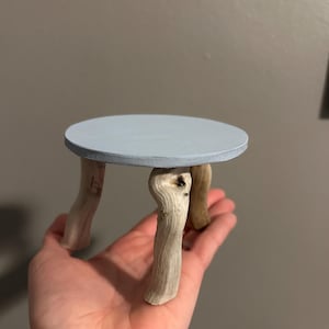 Puede incluir: Una pequeña mesa de madera hecha a mano con una tapa redonda pintada de azul claro. La mesa tiene tres patas hechas de madera flotante natural, mostrando la veta y textura únicas de la madera. La mesa tiene aproximadamente 10 cm de diámetro.