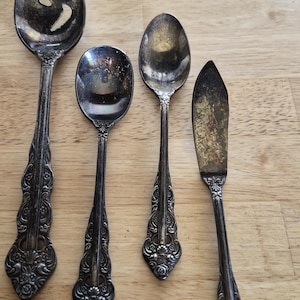 May include: A set of four tarnished silver-toned utensils featuring ornate, embossed floral handles. The collection includes a large slotted spoon, two smaller spoons of varying sizes, and a flat-edged butter knife with a pointed tip.
