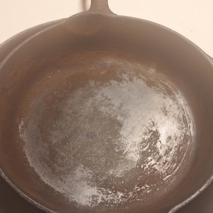 May include: Two cast iron skillets, one nested inside the other. The skillets are dark brown with a well-seasoned cooking surface. The top skillet has a handle with a hole for hanging. The background is a light beige.