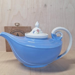 May include: A light blue ceramic teapot with a white handle and lid. The lid is decorated with floral patterns. The teapot is on a wooden surface. The Etsy shop URL is visible in the image.