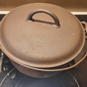 May include: A brown cast iron Dutch oven with a lid and handle, sitting on a black stovetop. The oven has a rounded shape and a metal handle on the lid. The stovetop has control knobs and a white surface.