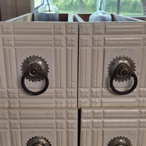 May include: Two stacked off-white wooden drawer units featuring a grid-like embossed pattern on the front panels. Each drawer is equipped with a circular, dark metal ring pull handle attached to a decorative, scalloped, antique-style metal backplate.