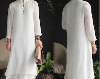 May include: A white, long-sleeved dress with a mandarin collar and a layered skirt. The dress is made of a lightweight fabric and has a relaxed fit. The model is wearing brown sandals and holding a rolled-up paper.