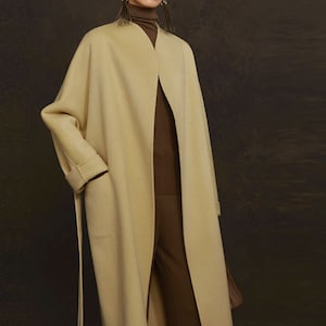 Könnte beinhalten: Ein langer, beiger Wollmantel mit einem Selbstgürtel und einem schlichten, minimalistischen Design.