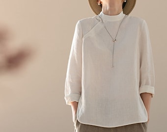 May include: A white linen top with a mandarin collar and three-quarter sleeves, paired with wide-leg, tan linen pants. The top features a unique asymmetrical button closure and a delicate necklace. The outfit is simple and elegant.