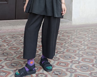 Puede incluir: Un top negro holgado de manga corta con cintura fruncida y un bolso tote negro. La persona lleva pantalones negros de pierna ancha y calcetines coloridos con sandalias negras.