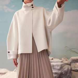 May include: A white oversized collared jacket with a single button closure and long sleeves. The jacket has a slightly asymmetrical hemline and is paired with a pleated skirt.