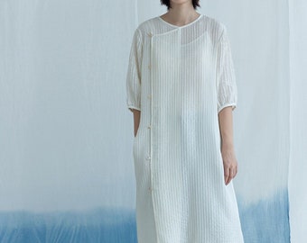 May include: A white, vertically striped dress with a button-up front and short sleeves. The dress is paired with dark denim cropped pants and dark shoes. The background is a gradient of white and blue.
