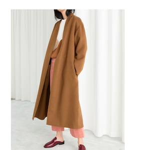 Könnte beinhalten: Ein langer brauner Mantel mit Schalkragen und lockerer Passform. Der Mantel wird über einem weißen Pullover und einer rosa Hose getragen. Die Person trägt burgunderfarbene Leder-Slipper.